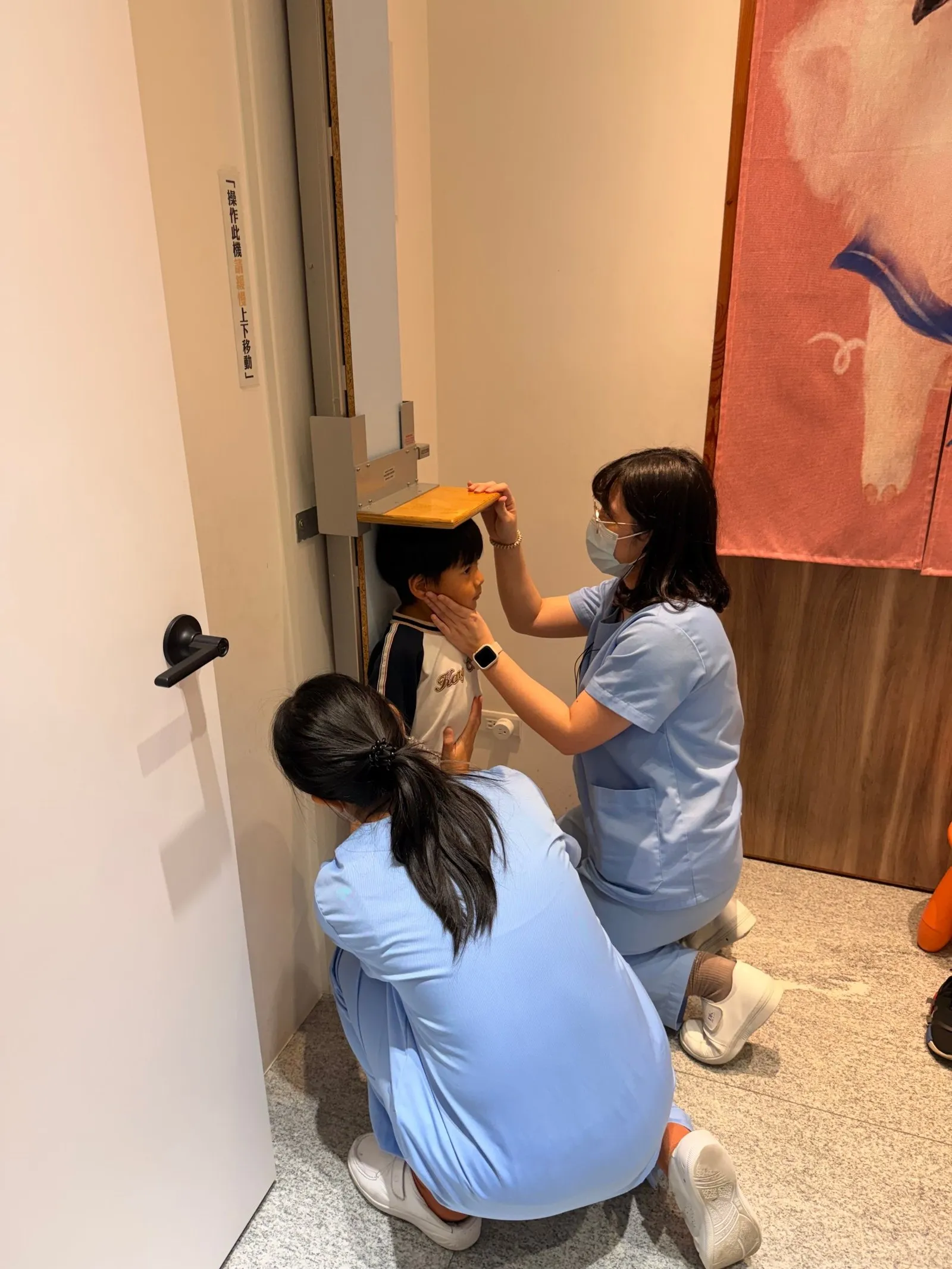 兩位護理師一起量身高:一位扶著下巴讓頭擺正,一位蹲著扶身體貼牆,頭頂的木板慢慢壓下來