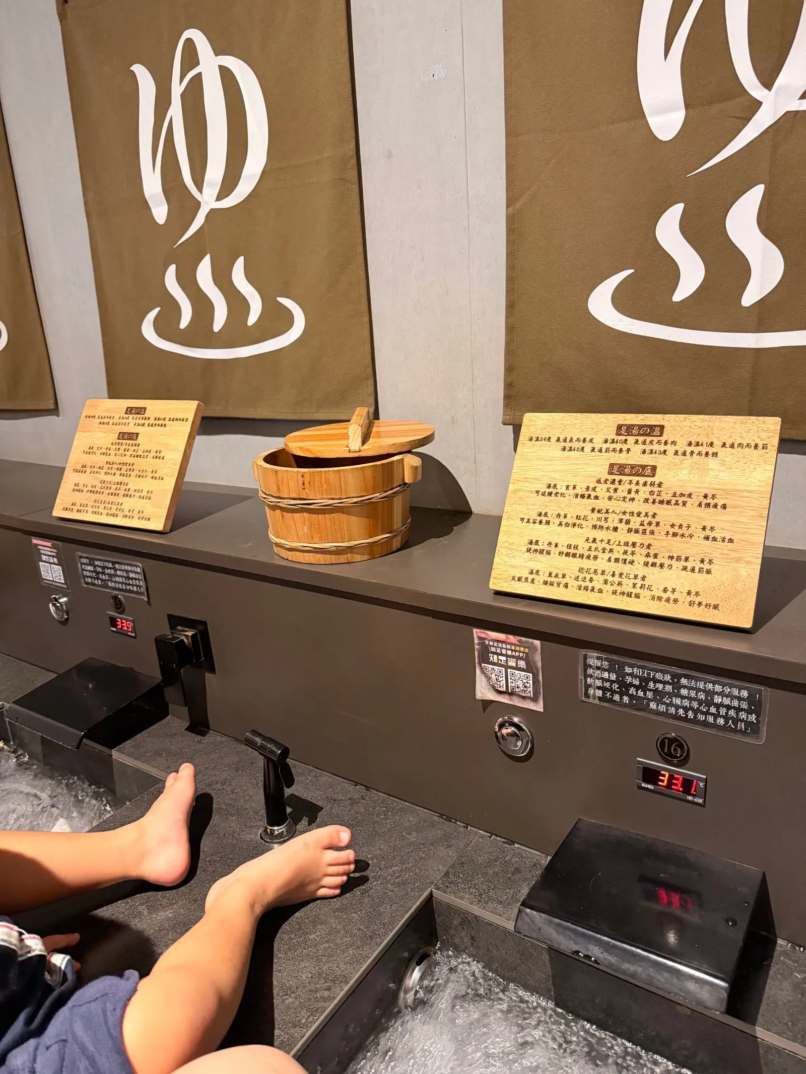 一整排泡腳池,上方掛著「ゆ」字布簾,檯面擺著木桶與介紹湯溫、湯底的木牌,池邊數字顯示 33.1°C,兩雙腳泡在水裡