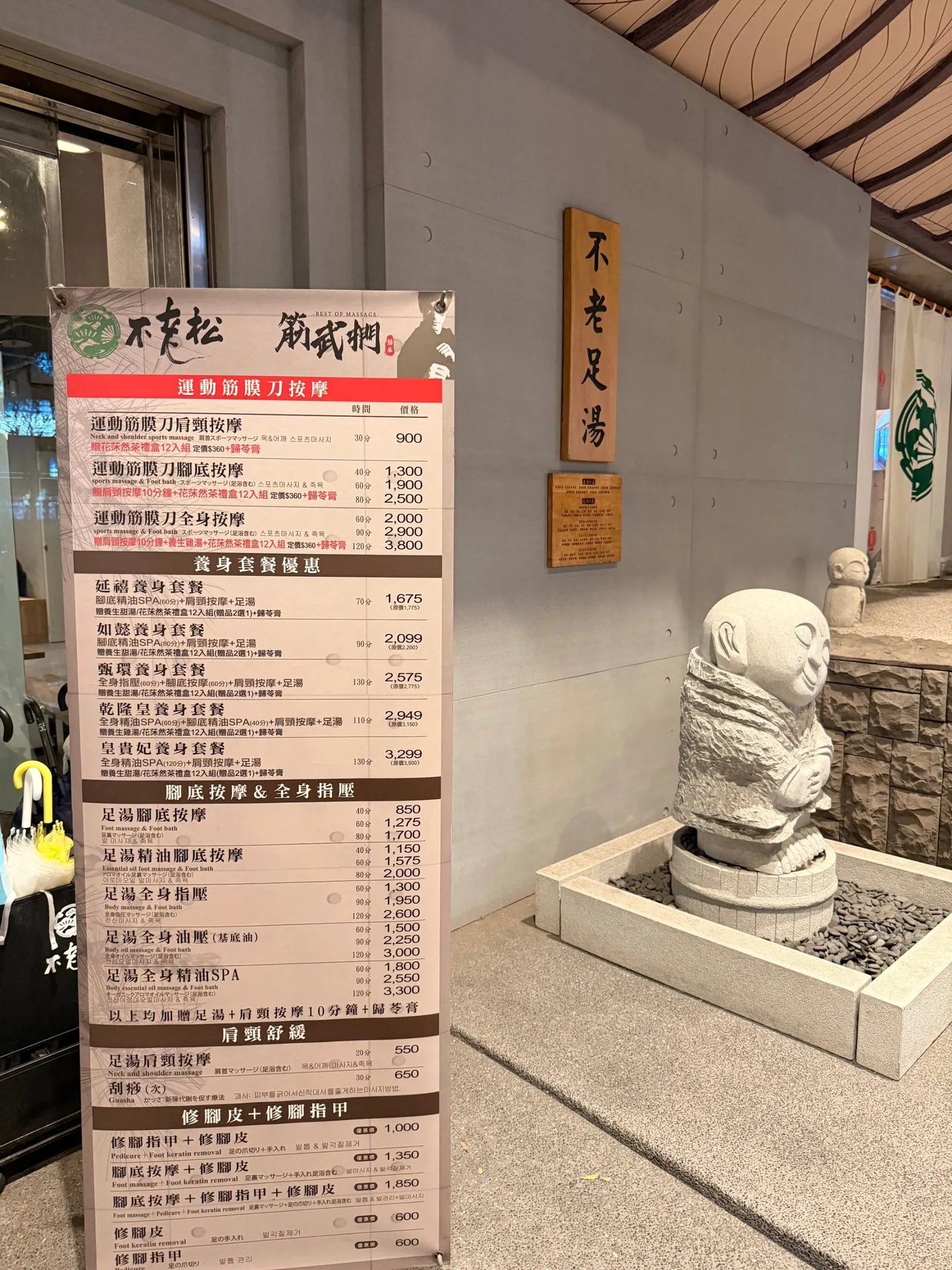 不老松門口的立式價目看板,分成運動筋膜刀按摩、養身套餐優惠、腳底按摩與全身指壓、肩頸舒緩、修腳皮等區塊,旁邊是「不老足湯」木牌與石雕